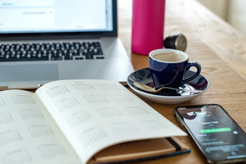 Planner and phone in front of a laptop and a cup of coffee